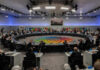 A large round table meeting with global leaders and flags in the background