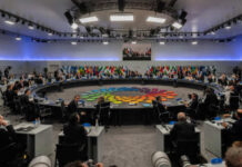 A large round table meeting with global leaders and flags in the background
