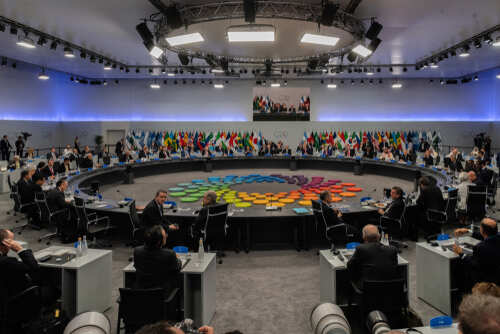 A large round table meeting with global leaders and flags in the background