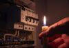 A hand holding a lit candle near an electrical panel
