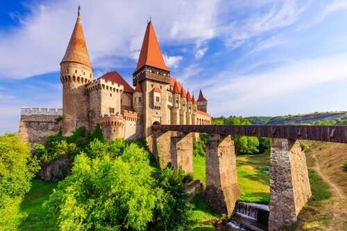 shutterstock_2445219915.jpg A medieval castle with tall towers and a wooden bridge surrounded by greenery