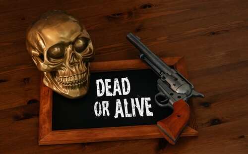 shutterstock_2433421607.jpg A golden skull next to a revolver on a wooden table with a sign saying 'DEAD OR ALIVE'
