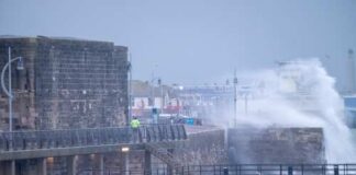 A man in a yellow jacket walking along a pier during a storm with crashing waves
