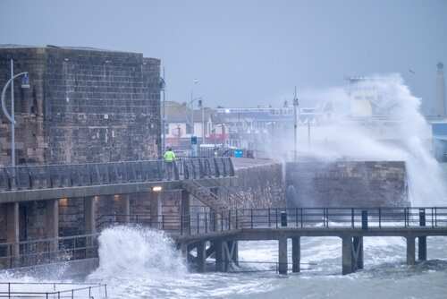 shutterstock_2575938495.jpg A man in a yellow jacket walking along a pier during a storm with crashing waves