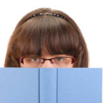 A girl with glasses peeking over a blue book