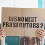A person holding a cardboard sign that reads 'DISHONEST PROSECUTORS'