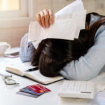 Person with head down on a desk surrounded by papers and credit cards