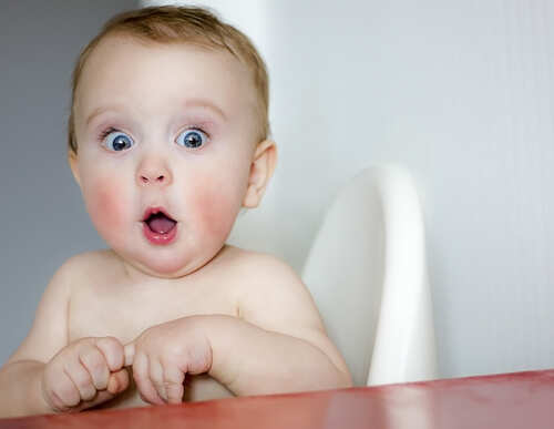 shutterstock_464300804.jpg A surprised baby sitting in a high chair with wide eyes and an open mouth