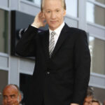 Maher Exposes Left’s “Never Enough” A man in a black suit smiling and gesturing at a public event