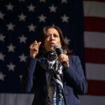 Kamala Harris Slams Trump’s ‘Dangerous Gamble’ Person speaking into microphone in front of U.S. flag.