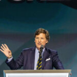 Tucker EXPLODES — Calls Trump Strikes Pure Evil Man speaking at podium with microphone and gesturing