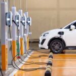 EV Giant Slashes Workforce A white electric vehicle parked at a charging station with multiple chargers