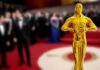 Golden Oscar statue in the foreground with blurred celebrities in formal attire in the background