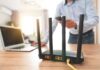 FCC Bans Foreign-Made Wireless Routers: China’s Spying On Us? A Wi-Fi router with antennas on a wooden desk next to a laptop and a person using a smartphone