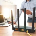 A Wi-Fi router with antennas on a wooden desk next to a laptop and a person using a smartphone