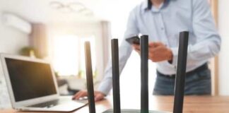 A Wi-Fi router with antennas on a wooden desk next to a laptop and a person using a smartphone