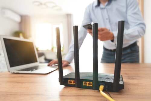 A Wi-Fi router with antennas on a wooden desk next to a laptop and a person using a smartphone