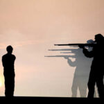 Silhouette of a person standing before soldiers aiming rifles at sunset