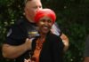 A smiling woman in a bright orange headscarf and necklace at an outdoor event with a police officer in the background