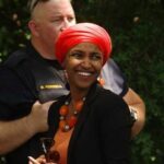 A smiling woman in a bright orange headscarf and necklace at an outdoor event with a police officer in the background