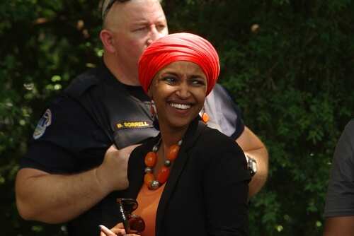 A smiling woman in a bright orange headscarf and necklace at an outdoor event with a police officer in the background
