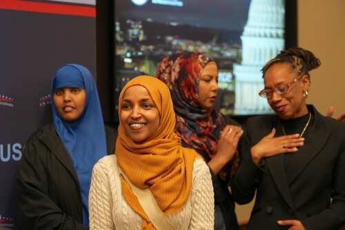 A group of women in cultural attire smiling at an event with a political backdrop