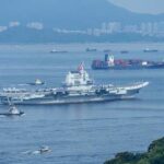 A naval ship navigating through the ocean with cargo ships and a city skyline in the background