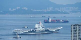 A naval ship navigating through the ocean with cargo ships and a city skyline in the background