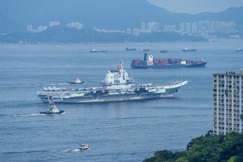 A naval ship navigating through the ocean with cargo ships and a city skyline in the background