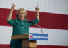 Hillary Stuns: Praises Trump’s Plan Person speaking emphatically at podium against striped backdrop