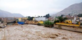 A flooded river flowing through an urban area with buildings in the background