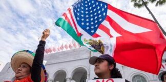 Two women at a protest holding flags and a sign against deportation