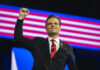 Oval Office Shock: Pratt Photo Ignites Firestorm A man in a suit raises his fist in a confident pose on stage with an American flag backdrop
