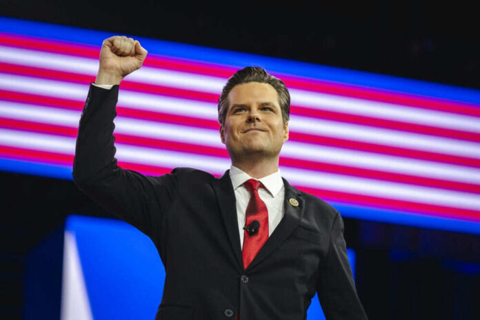A man in a suit raises his fist in a confident pose on stage with an American flag backdrop