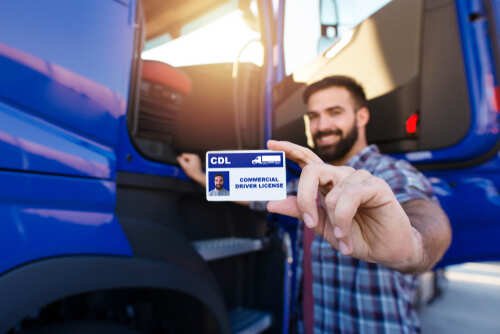 Portrait,Of,Middle,Aged,Bearded,Truck,Driver,Standing,By,The Man holding a commercial driver's license in front of a blue truck