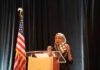 A woman in a hijab speaking passionately at a podium with an American flag in the background
