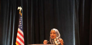 A woman in a hijab speaking passionately at a podium with an American flag in the background