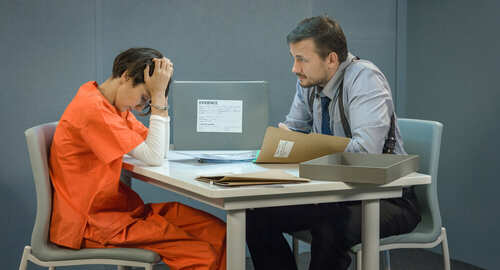 Detective,In,The,Interrogation,Room,With,A,Box,Of,Evidence A detective interrogating a prisoner in an orange jumpsuit at a table