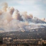 Human Spark, City Inferno A wildfire producing thick smoke over a city landscape with a helicopter in the sky