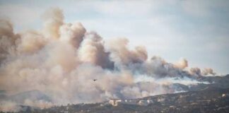 A wildfire producing thick smoke over a city landscape with a helicopter in the sky
