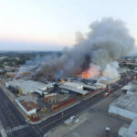 A large industrial fire with thick smoke rising into the sky and firefighters responding