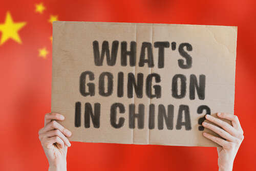 shutterstock_1812983458 Hands holding a cardboard sign with the text 'What's going on in China?' against a red background with stars