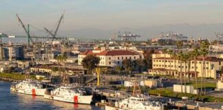 Three U.S. Coast Guard boats docked at a harbor with cranes in the background